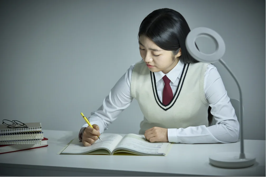 책상 조명 아래 펼친 책에 필기하는 학생