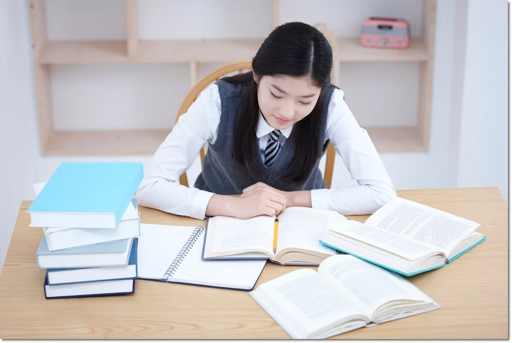 쌓인 책 옆 펼친 교재에 글을 쓰는 학생